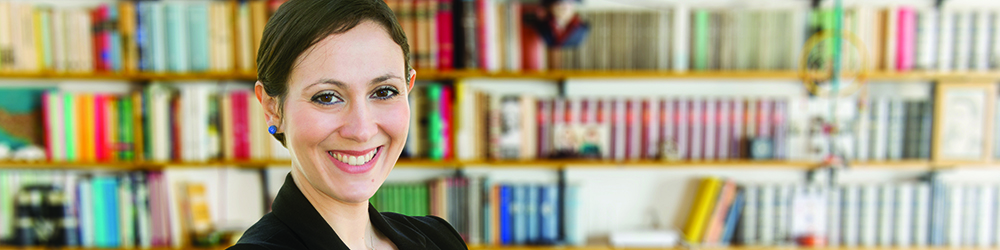 A woman in a suit stands in front of a crowded bookshelf