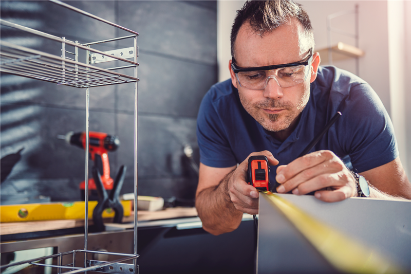 Gentleman wearing safety glasses using a tape measure to measure the dimensions of a shelf