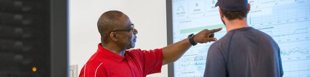 A man points at a large screen displaying charts and graphs as another man looks on.