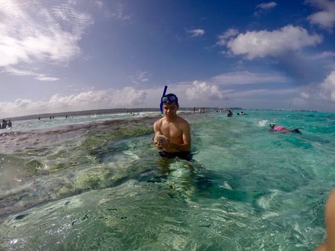A young man wearing goggles and a snorkle on his forehead looks at a seacreature he has found in waist-deep waters.