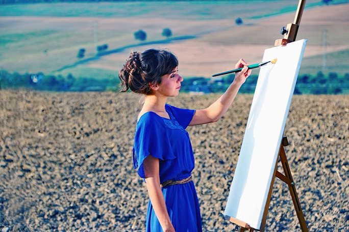 A woman in a blue dress paints standing up in a field in the countryside.