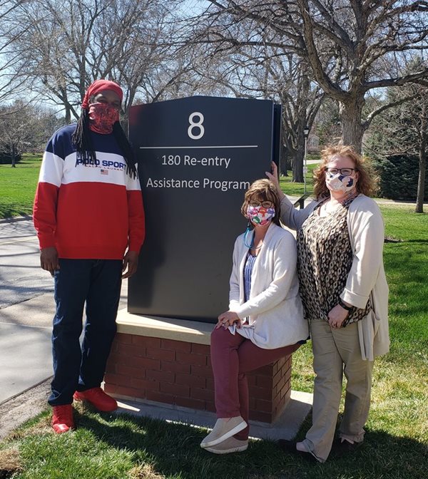 Three people pose in front of the sign for Building 8 on Fort Omaha Campus, home of 180 Re-entry Assistance Program